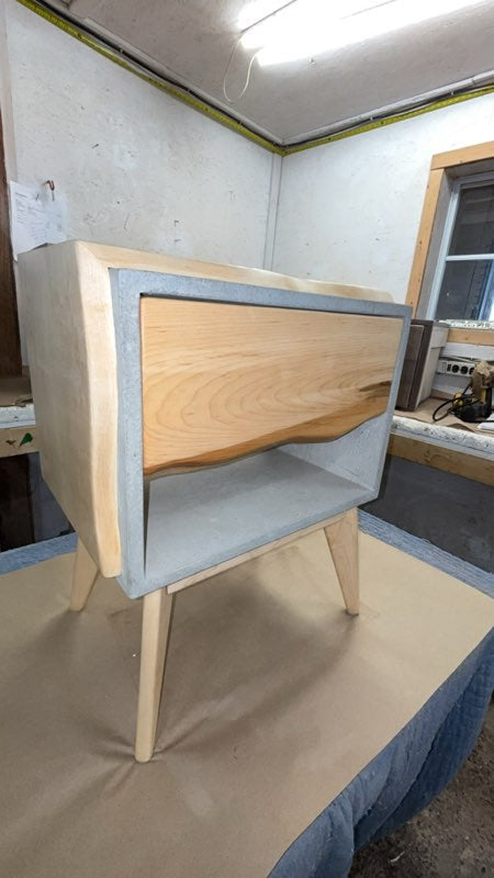 A mid-century modern wooden bedside table with a maple solid wood drawer front, a recessed open shelf, and tapered legs, photographed inside a workshop or studio.